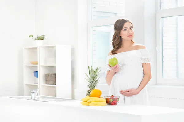 Bella donna incinta sorridente in cucina mangiare frutta — Foto Stock