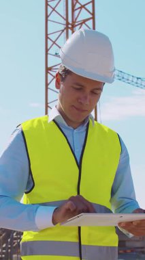 İnşaat alanının önünde duran profesyonel müteahhit. Kasklı ve yelekli ustabaşı. Ofis binası ve vinç arka planı. İş, gayrimenkul ve yatırım kavramları.