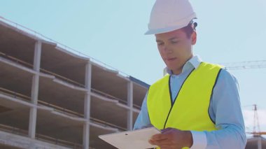 İnşaat alanının önünde duran profesyonel müteahhit. Kasklı ve yelekli ustabaşı. Ofis binası ve vinç arka planı. İş, gayrimenkul ve yatırım kavramları.