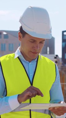 İnşaat alanının önünde duran profesyonel müteahhit. Kasklı ve yelekli ustabaşı. Ofis binası ve vinç arka planı. İş, gayrimenkul ve yatırım kavramları.