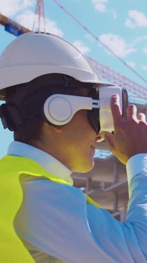 İnşaat alanının önünde duran profesyonel müteahhit. Kasklı ve yelekli ustabaşı. Ofis binası ve vinç arka planı. İş, gayrimenkul ve yatırım kavramları.