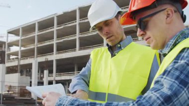 Büyük bir inşaat sahasının mimari planlarını inceleyen sert şapka ve güvenlik yelekleri giyen profesyonel erkek mühendisler. Odaklanmış inşaat işçileri ve site yöneticileri proje planları ve yapısal gelişimi gün boyunca tartıştılar