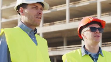 Büyük bir inşaat sahasının mimari planlarını inceleyen sert şapka ve güvenlik yelekleri giyen profesyonel erkek mühendisler. Odaklanmış inşaat işçileri ve site yöneticileri proje planları ve yapısal gelişimi gün boyunca tartıştılar