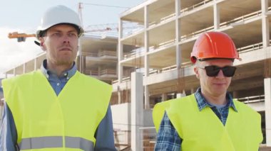 Büyük bir inşaat sahasının mimari planlarını inceleyen sert şapka ve güvenlik yelekleri giyen profesyonel erkek mühendisler. Odaklanmış inşaat işçileri ve site yöneticileri proje planları ve yapısal gelişimi gün boyunca tartıştılar