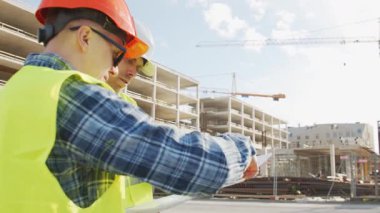 Büyük bir inşaat sahasının mimari planlarını inceleyen sert şapka ve güvenlik yelekleri giyen profesyonel erkek mühendisler. Odaklanmış inşaat işçileri ve site yöneticileri proje planları ve yapısal gelişimi gün boyunca tartıştılar