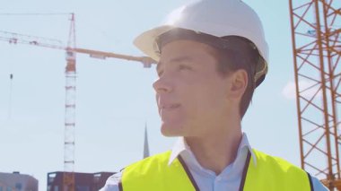 İnşaat alanının önünde duran profesyonel müteahhit. Kasklı ve yelekli ustabaşı. Ofis binası ve vinç arka planı. İş, gayrimenkul ve yatırım kavramları.