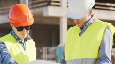 Büyük bir inşaat sahasının mimari planlarını inceleyen sert şapka ve güvenlik yelekleri giyen profesyonel erkek mühendisler. Odaklanmış inşaat işçileri ve site yöneticileri proje planları ve yapısal gelişimi gün boyunca tartıştılar