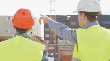 Büyük bir inşaat sahasının mimari planlarını inceleyen sert şapka ve güvenlik yelekleri giyen profesyonel erkek mühendisler. Odaklanmış inşaat işçileri ve site yöneticileri proje planları ve yapısal gelişimi gün boyunca tartıştılar