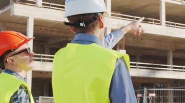 Büyük bir inşaat sahasının mimari planlarını inceleyen sert şapka ve güvenlik yelekleri giyen profesyonel erkek mühendisler. Odaklanmış inşaat işçileri ve site yöneticileri proje planları ve yapısal gelişimi gün boyunca tartıştılar