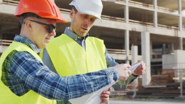 Büyük bir inşaat sahasının mimari planlarını inceleyen sert şapka ve güvenlik yelekleri giyen profesyonel erkek mühendisler. Odaklanmış inşaat işçileri ve site yöneticileri proje planları ve yapısal gelişimi gün boyunca tartıştılar