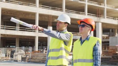 Büyük bir inşaat sahasının mimari planlarını inceleyen sert şapka ve güvenlik yelekleri giyen profesyonel erkek mühendisler. Odaklanmış inşaat işçileri ve site yöneticileri proje planları ve yapısal gelişimi gün boyunca tartıştılar