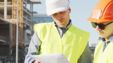 Büyük bir inşaat sahasının mimari planlarını inceleyen sert şapka ve güvenlik yelekleri giyen profesyonel erkek mühendisler. Odaklanmış inşaat işçileri ve site yöneticileri proje planları ve yapısal gelişimi gün boyunca tartıştılar