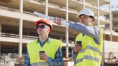 Büyük bir inşaat sahasının mimari planlarını inceleyen sert şapka ve güvenlik yelekleri giyen profesyonel erkek mühendisler. Odaklanmış inşaat işçileri ve site yöneticileri proje planları ve yapısal gelişimi gün boyunca tartıştılar
