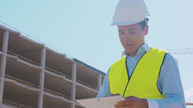 İnşaat alanının önünde duran profesyonel müteahhit. Kasklı ve yelekli ustabaşı. Ofis binası ve vinç arka planı. İş, gayrimenkul ve yatırım kavramları.