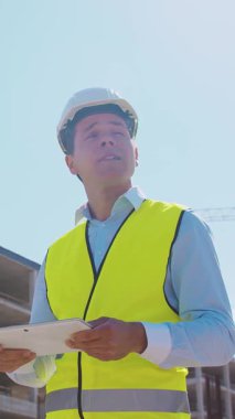 İnşaat alanının önünde duran profesyonel müteahhit. Kasklı ve yelekli ustabaşı. Ofis binası ve vinç arka planı. İş, gayrimenkul ve yatırım kavramları.