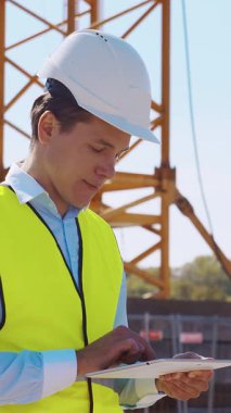 İnşaat alanının önünde duran profesyonel müteahhit. Kasklı ve yelekli ustabaşı. Ofis binası ve vinç arka planı. İş, gayrimenkul ve yatırım kavramları.