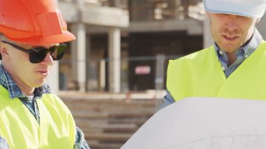 Büyük bir inşaat sahasının mimari planlarını inceleyen sert şapka ve güvenlik yelekleri giyen profesyonel erkek mühendisler. Odaklanmış inşaat işçileri ve site yöneticileri proje planları ve yapısal gelişimi gün boyunca tartıştılar