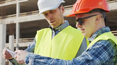 Büyük bir inşaat sahasının mimari planlarını inceleyen sert şapka ve güvenlik yelekleri giyen profesyonel erkek mühendisler. Odaklanmış inşaat işçileri ve site yöneticileri proje planları ve yapısal gelişimi gün boyunca tartıştılar