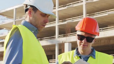 Büyük bir inşaat sahasının mimari planlarını inceleyen sert şapka ve güvenlik yelekleri giyen profesyonel erkek mühendisler. Odaklanmış inşaat işçileri ve site yöneticileri proje planları ve yapısal gelişimi gün boyunca tartıştılar