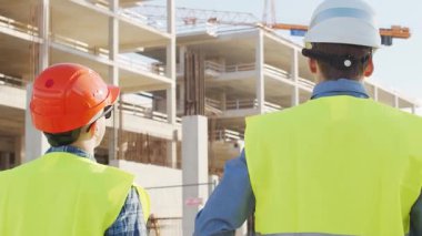 Büyük bir inşaat sahasının mimari planlarını inceleyen sert şapka ve güvenlik yelekleri giyen profesyonel erkek mühendisler. Odaklanmış inşaat işçileri ve site yöneticileri proje planları ve yapısal gelişimi gün boyunca tartıştılar