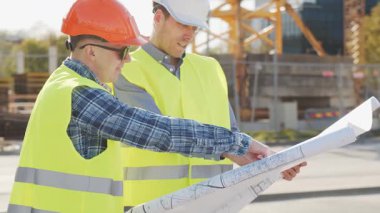 Büyük bir inşaat sahasının mimari planlarını inceleyen sert şapka ve güvenlik yelekleri giyen profesyonel erkek mühendisler. Odaklanmış inşaat işçileri ve site yöneticileri proje planları ve yapısal gelişimi gün boyunca tartıştılar