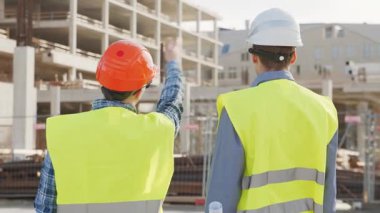 Büyük bir inşaat sahasının mimari planlarını inceleyen sert şapka ve güvenlik yelekleri giyen profesyonel erkek mühendisler. Odaklanmış inşaat işçileri ve site yöneticileri proje planları ve yapısal gelişimi gün boyunca tartıştılar