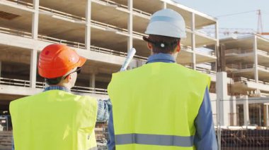 Büyük bir inşaat sahasının mimari planlarını inceleyen sert şapka ve güvenlik yelekleri giyen profesyonel erkek mühendisler. Odaklanmış inşaat işçileri ve site yöneticileri proje planları ve yapısal gelişimi gün boyunca tartıştılar