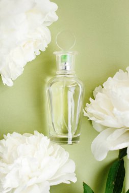 Parfümlü şeffaf model cam şişe zeytin yeşili pastel renginde, taze beyaz şakayık çiçekleri arasında.