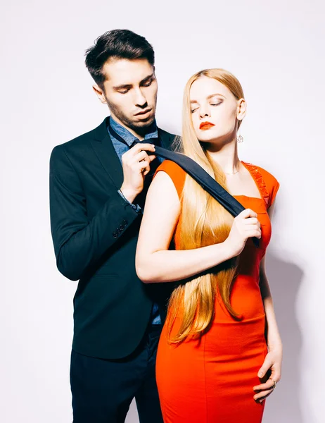 Young fashion man and woman against white wall, posing for the camera. Woman pulls man for a tie. Indoor. Warm color. — Stock Photo, Image