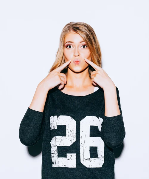 Beautiful young blond girl posing in Sweatshirt and white shorts and red lips kiss does. She points a finger at her lips. Indoor. Close up. Warm color — Stock Photo, Image