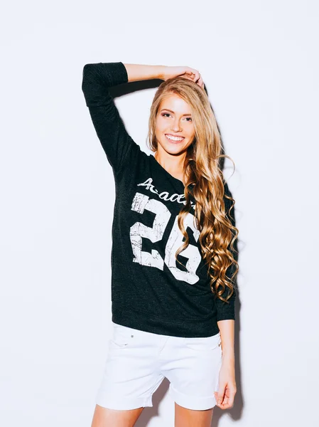 Muy hermosa chica con pelo largo Rubio posando sobre un fondo blanco. Ella levantó su mano sobre su cabeza y sonriendo. Shorts blanco y sudadera. Interior. Color caliente. — Foto de Stock