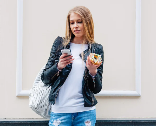 Retrato de una bella mujer sexy comiendo un donut, mira su teléfono inteligente en la calle ciudad europea. Al aire libre. — Foto de Stock