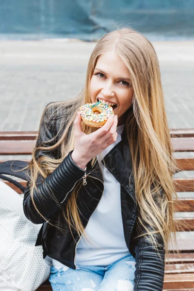 Atractivo joven sexy mujer rubia sentada en un banco en la Europa de la ciudad y comer sabroso buñuelo colorido. Retrato de estilo de vida al aire libre de niña bonita — Foto de Stock