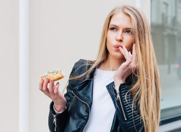 Hermosa mujer sexy joven comiendo un donut, lamiendo sus dedos tomando placer en una calle de ciudad europea. Al aire libre. Color caliente. Imágenes De Stock Sin Royalties Gratis