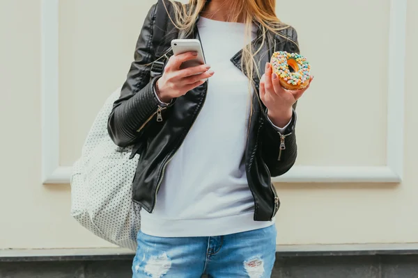 먹는 도넛, 아름 다운 젊은 섹시 한 여자의 초상화 거리 유럽 도시에 그녀의 스마트 폰에서 보인다. 바디 부분입니다. 야외. 클로즈업. 스톡 사진