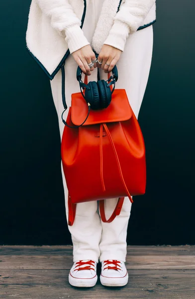 Moda mochila roja grande hermoso y auriculares en el brazo de la niña en un traje deportivo blanco moda e instructores, posando junto a la pared en una noche de verano. Color cálido. Cierre para arriba — Foto de Stock