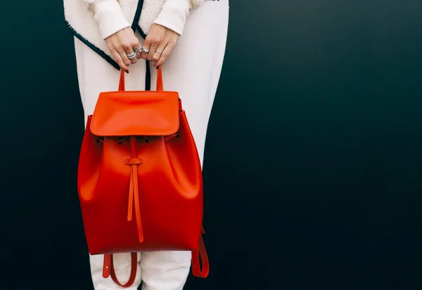 Fashionable beautiful big red backpack on the arm of the girl in a fashionable white sports suit, posing near the wall on a warm summer night. Part of the body. Warm color. Close up. — Stock Photo, Image