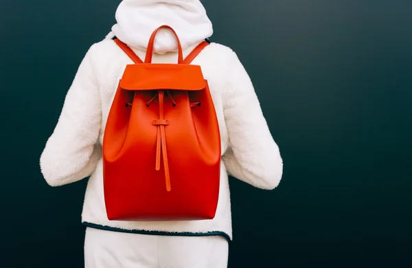 Fashionable beautiful big red backpack on the arm of the girl in a fashionable white sports suit, posing is back near the wall on a warm summer night. Part of the body. Warm color. Close up. - Stock-foto