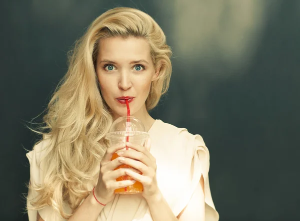 Beautiful sexy blonde with blue eyes drinking beverage through a straw on a hot summer day near the wall. Toned in warm colors — Stock Photo, Image