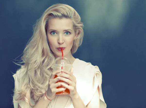 Beautiful sexy blonde with blue eyes drinking beverage through a straw on a hot summer day near the wall. Toned in warm colors — Stock Photo, Image