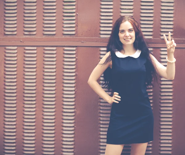 Beautiful brunette with long hair in a short black showing two fingers up near metallic brown fence. Colorful hipster photo — Stock Photo, Image