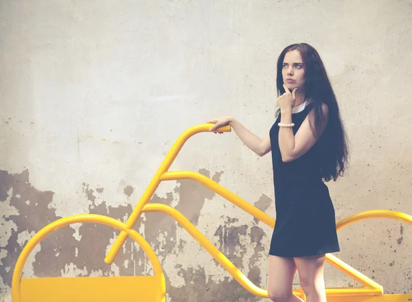 Beautiful brunette with long hair standing in thought near a yellow bike. Colorful hipster photo — Stock Photo, Image