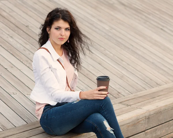 Beautiful tall girl with long hair brunette in jeans standing on old wooden planks with a cup of coffee in hand  on a warm summer evening — Stock Photo, Image