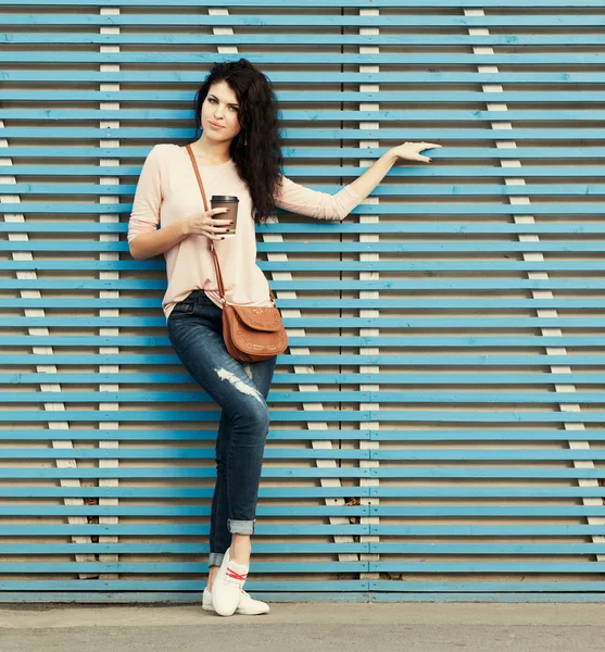 Mooie brunette meisje met een kopje koffie in de hand staat in de buurt van een muur van gekleurde houten planken — Stockfoto