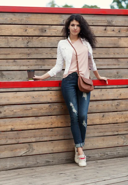 Belle grande fille aux cheveux longs brune en jeans debout sur de vieilles planches de bois avec une tasse de café à la main lors d'une chaude soirée d'été — Photo