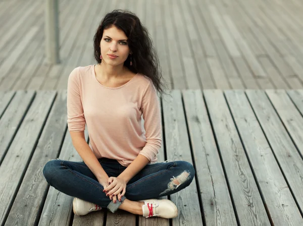 Mooi hoog meisje met lange haren brunette in spijkerbroek zit op houten planken — Stockfoto