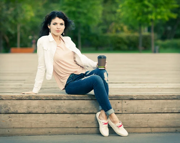 Hermosa chica alta con pelo largo morena en jeans sentados en tablones de madera viejos con una taza de café en la mano en una cálida noche de verano —  Fotos de Stock