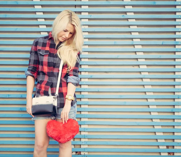Triste chica sosteniendo un corazón rojo y se encuentra cerca de una pared de tablones de madera colores — Zdjęcie stockowe