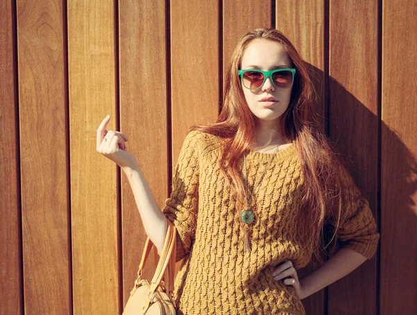 Bella ragazza rossa con grande borsa alla moda in occhiali da sole in piedi vicino alla parete di legno — Foto Stock