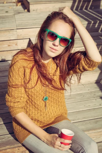 Menina ruiva bonita em óculos de sol sentado em tábuas de madeira em uma noite quente de verão — Fotografia de Stock